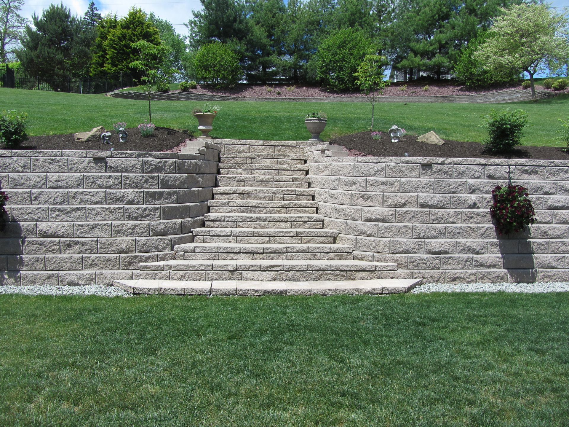 Stone steps leading up a grassy hill, flanked by tiered retaining walls and garden beds with small shrubs and planters.