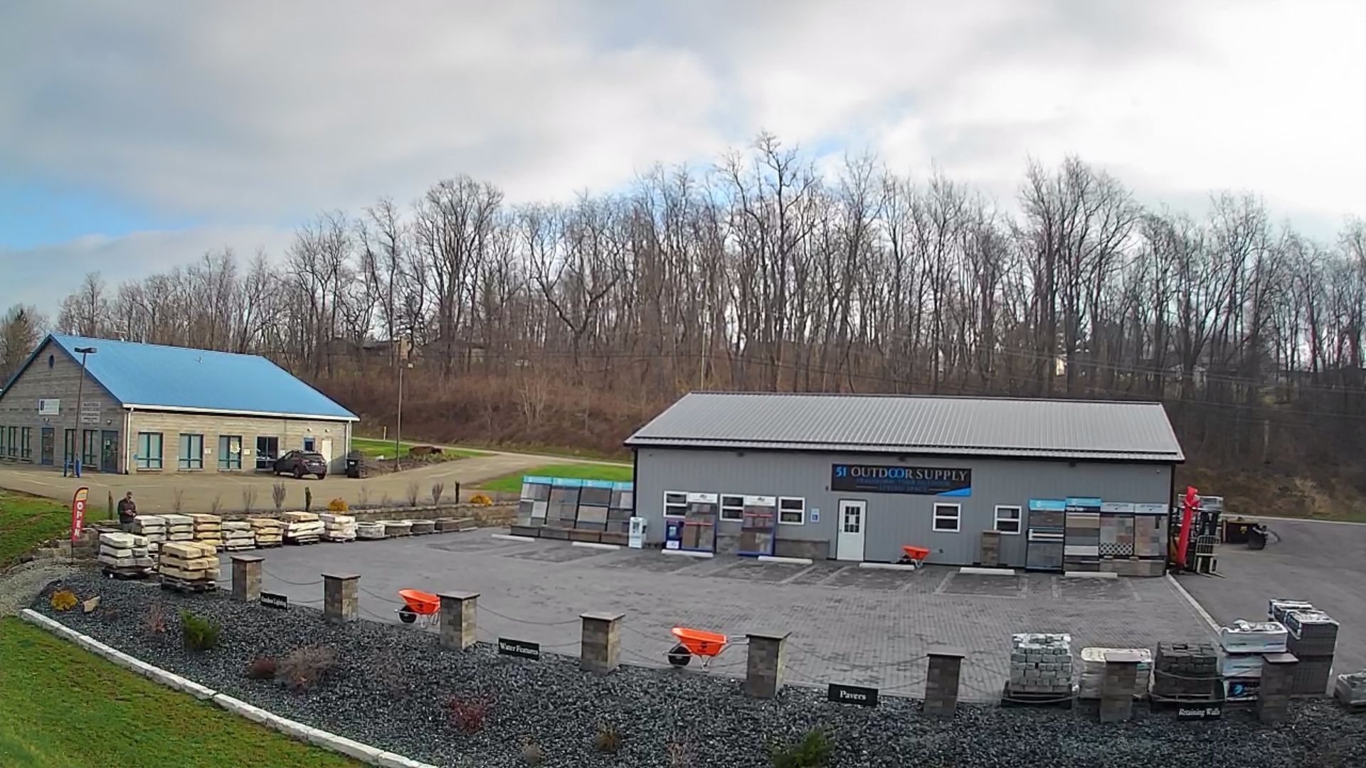 Two commercial buildings with grey exteriors, a blue roof, and a gravel parking lot lined with stone posts and equipment.