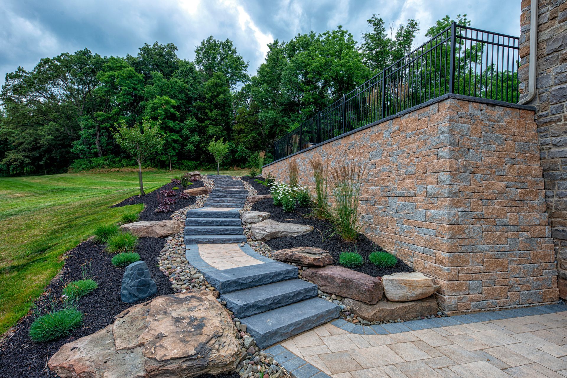 A stone staircase ascends through a landscaped garden next to a stacked stone retaining wall under a cloudy sky.
