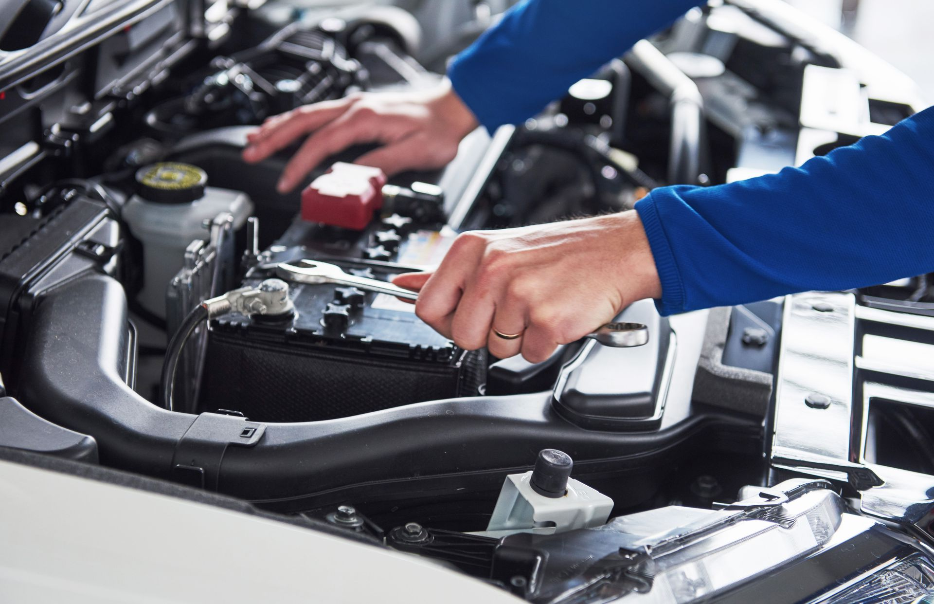 Hands working on a car engine, possibly checking the battery, in a well-lit environment.