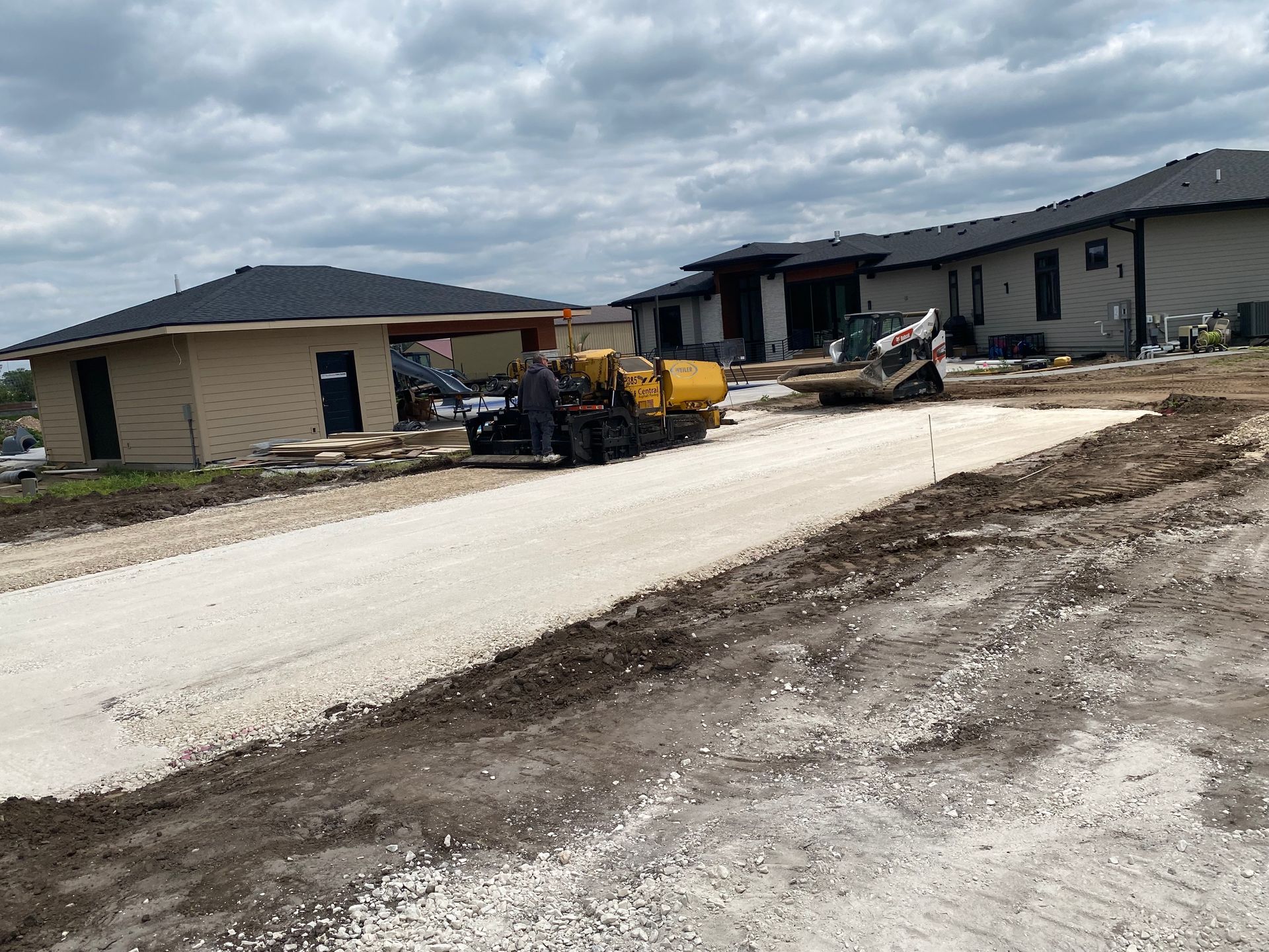 A small road under construction. A machine spreads gravel in front of a few houses under a cloudy sky.