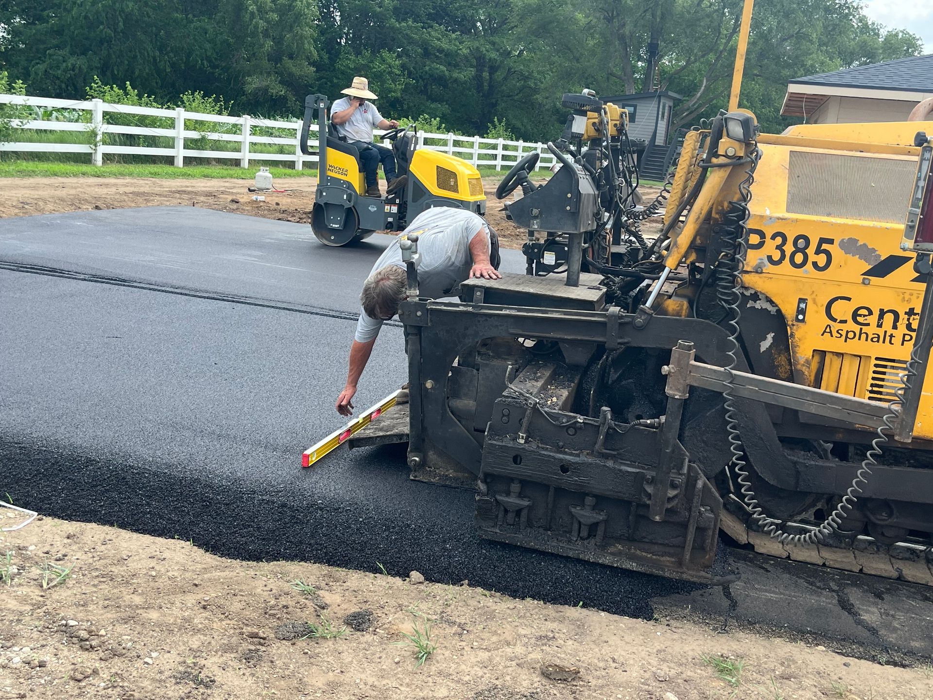 Road crew paving asphalt. Man measures asphalt level, other operates compactor and paver; setting is rural.