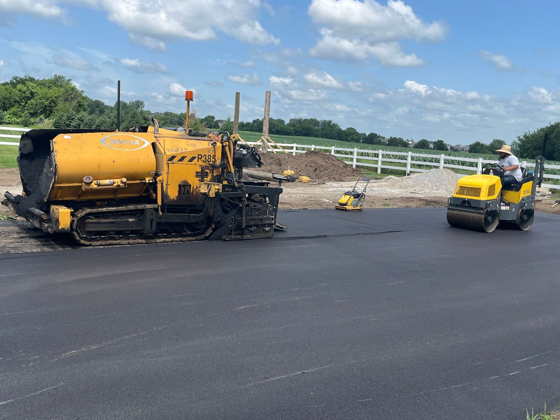 Asphalt paving in progress: a yellow paver and a compacting roller working on a new road surface.