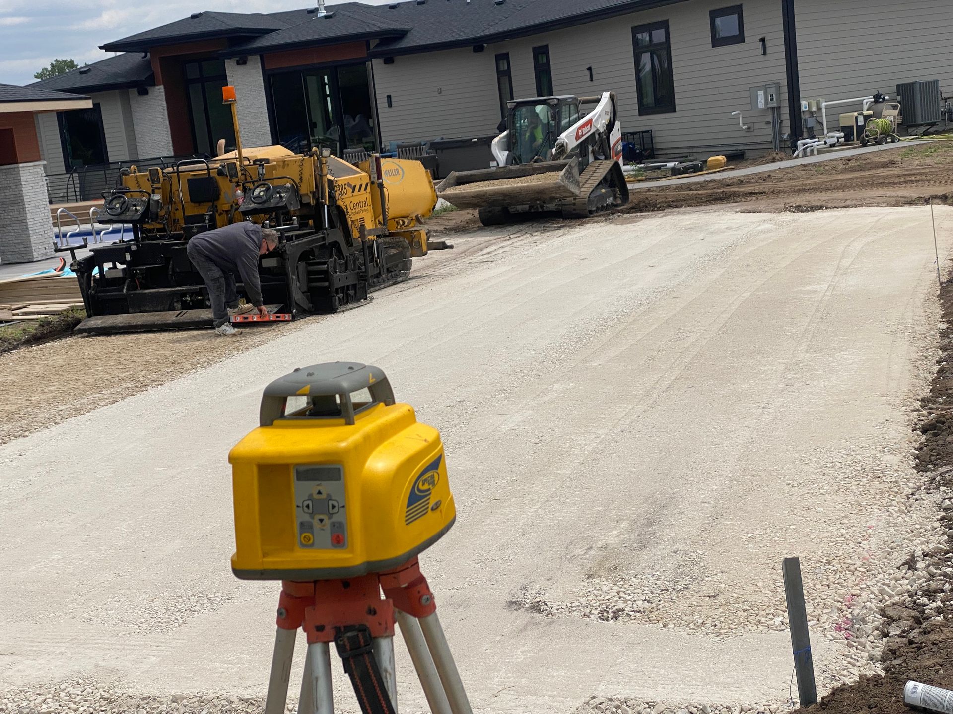 Yellow laser level on tripod; construction workers paving driveway with heavy machinery near a house.