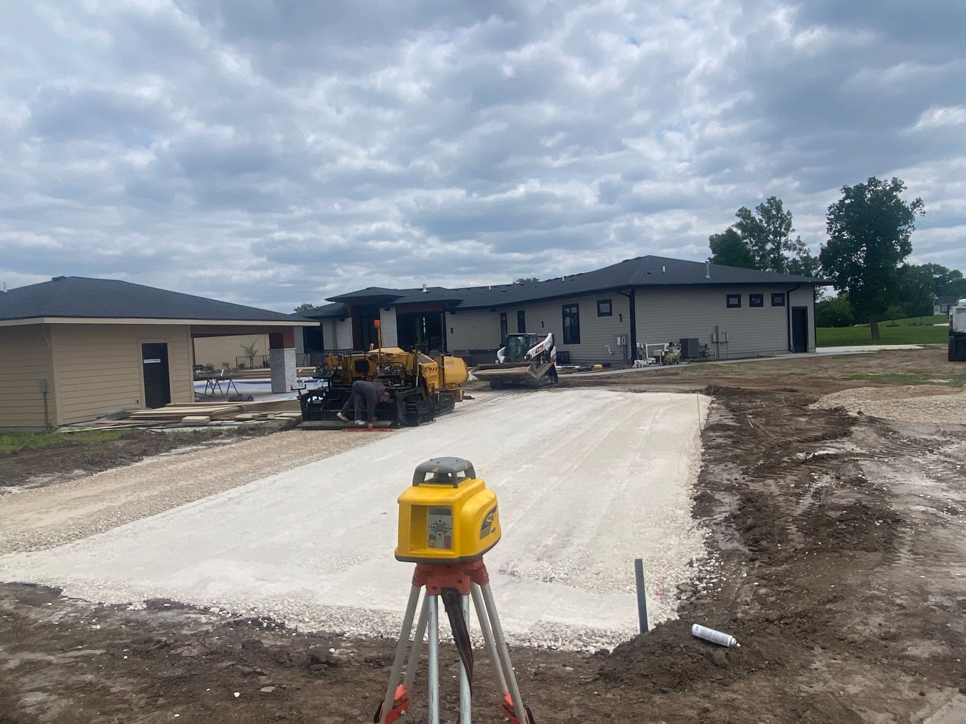 Construction site with gravel driveway; laser level in foreground, houses in background.