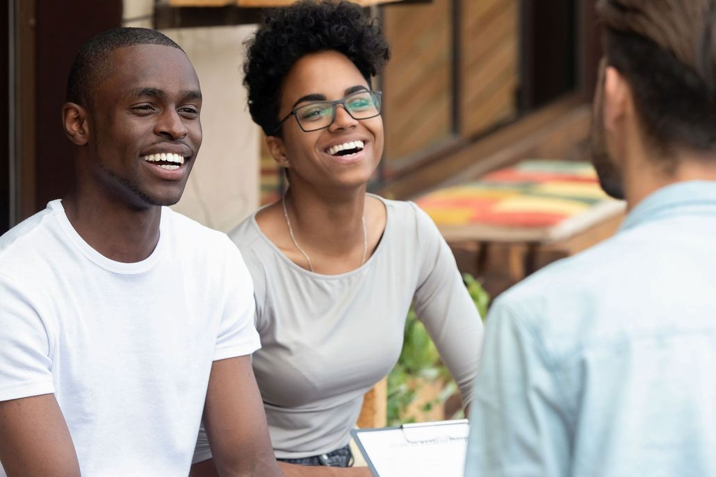 Two smiling people seated outdoors, talking with someone off-camera in a casual café setting