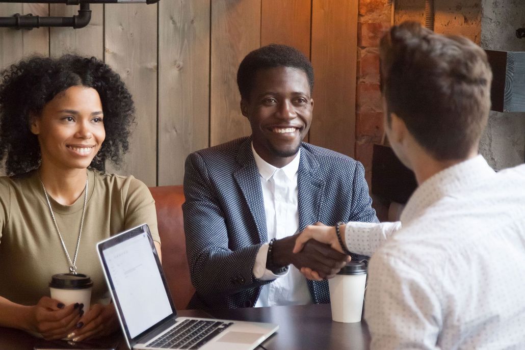 Three people in a meeting, shaking hands at a table with a laptop and coffee cups