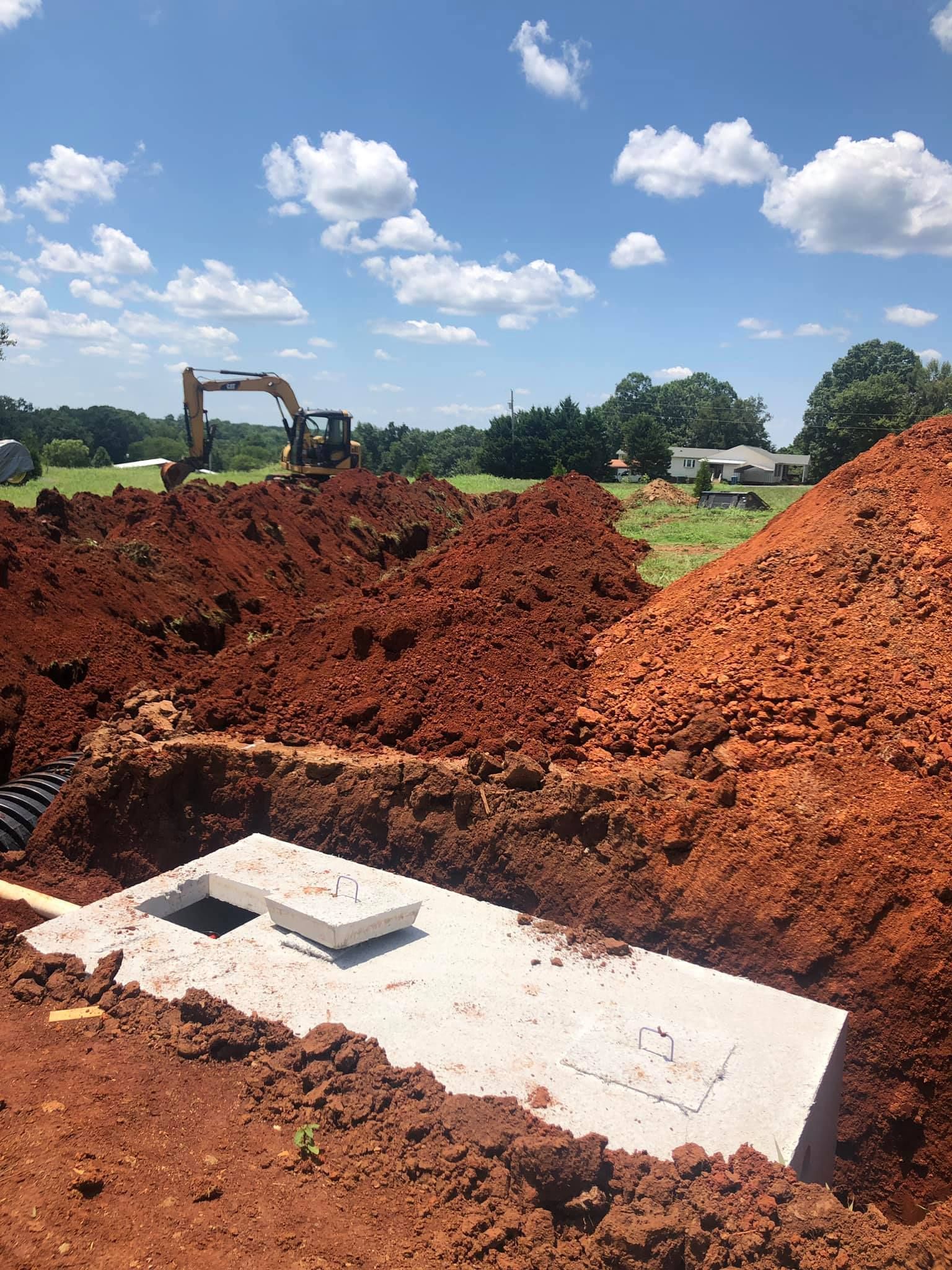 Concrete septic tank in a red-soil excavation with an excavator in the background under a blue sky.