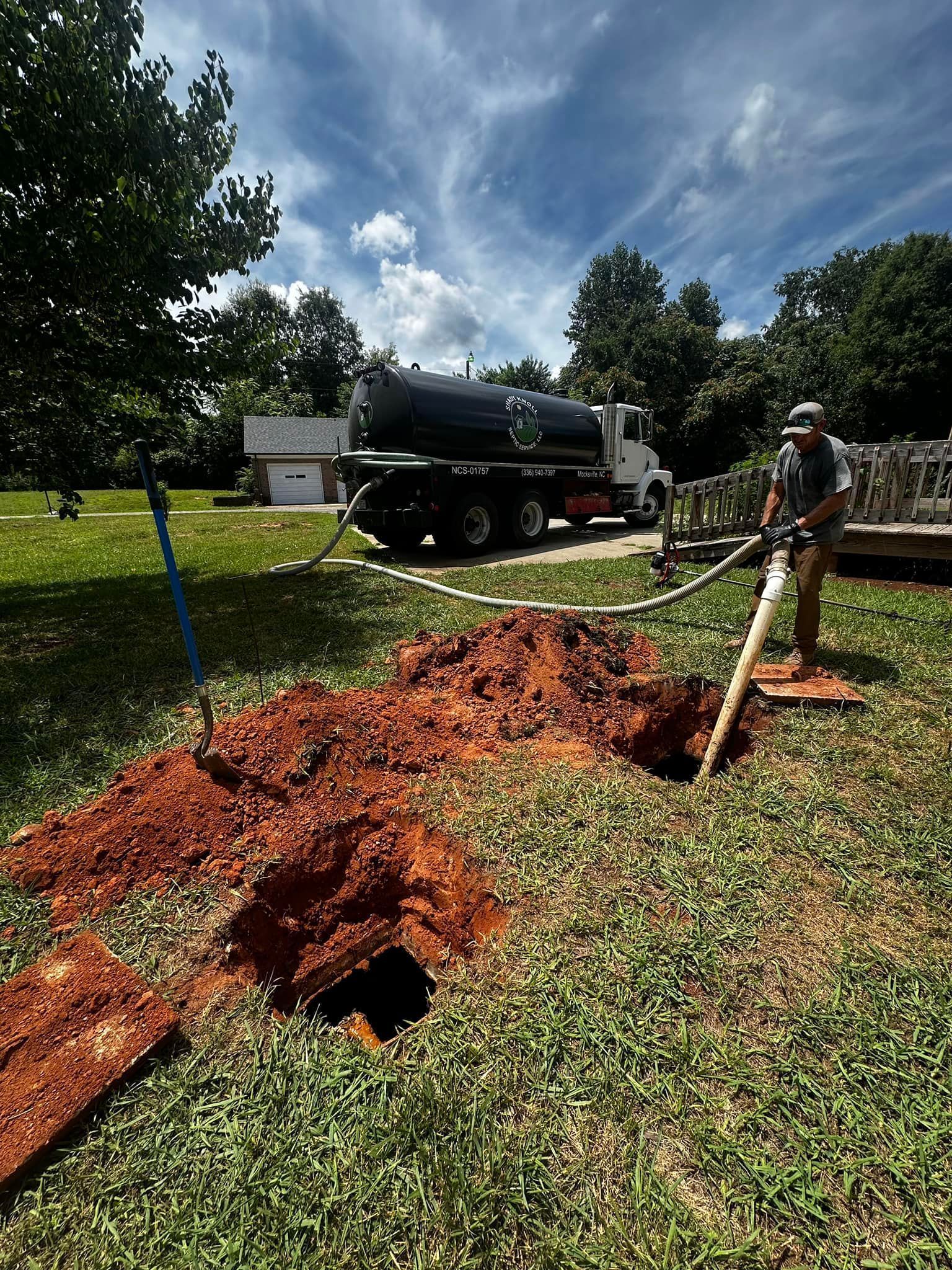 A worker pumping a septic tank. A truck is parked near an open ground hole on a sunny day.