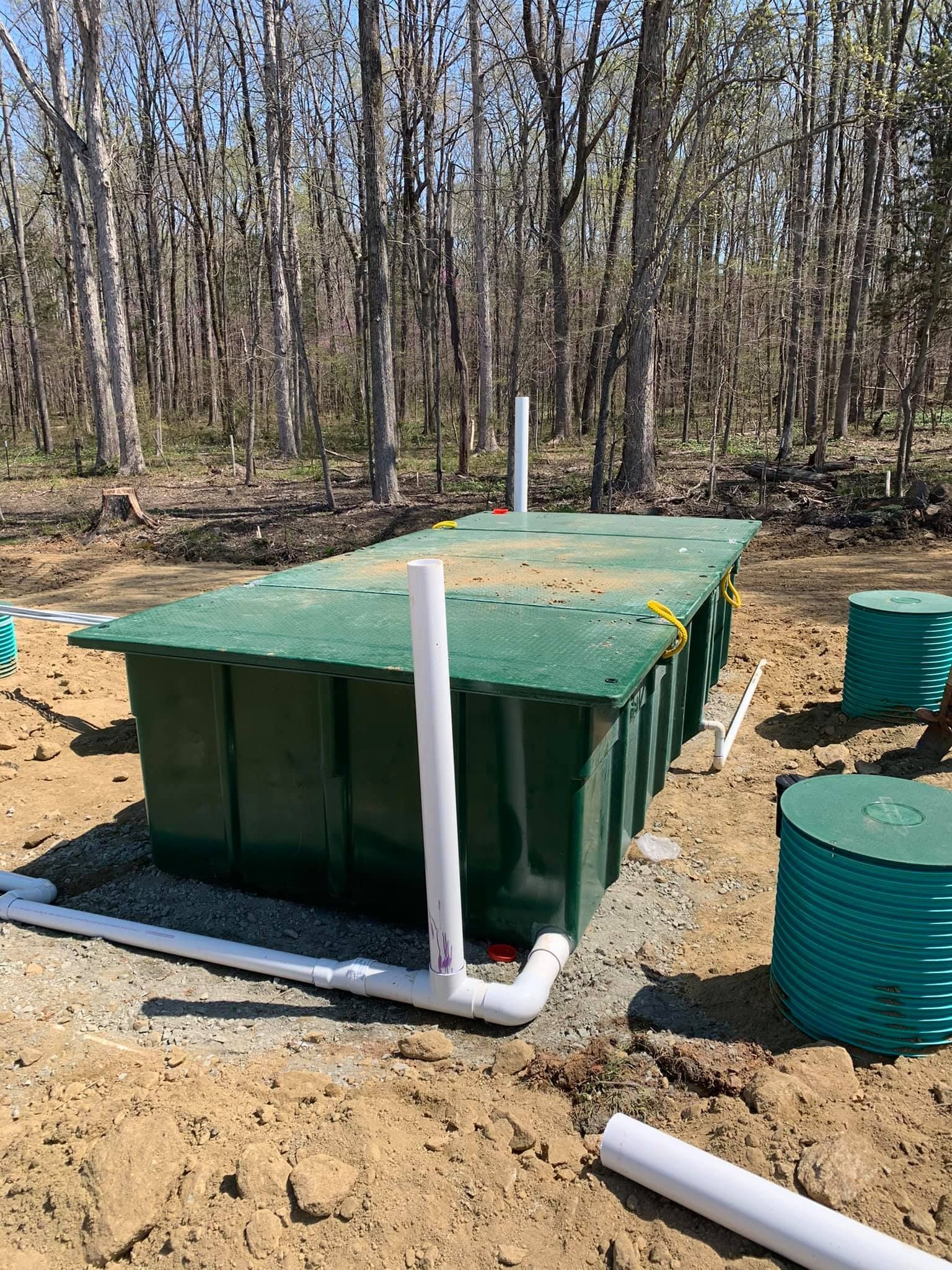Green septic tank with white pipes in a wooded area.