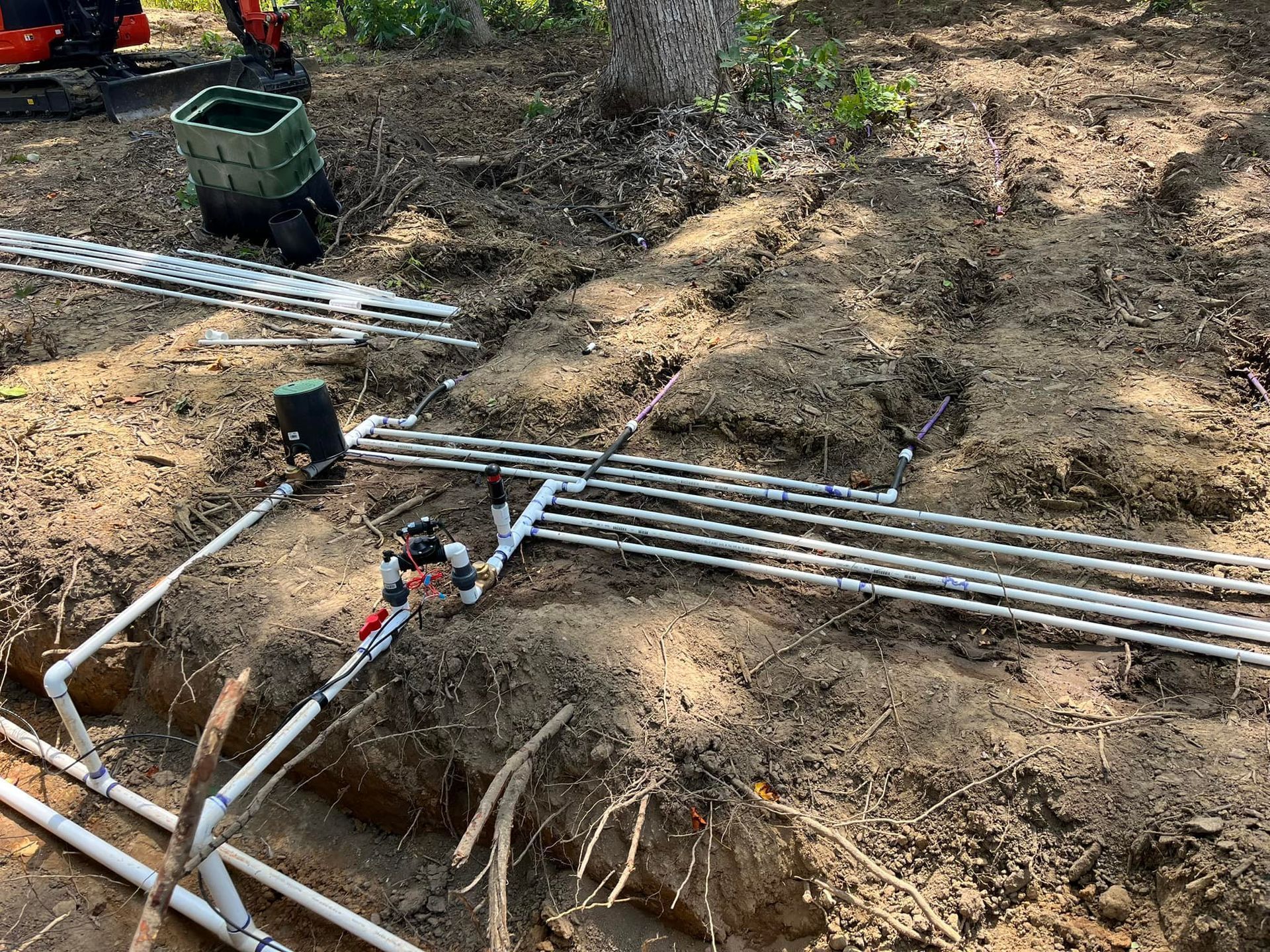 Irrigation system installation with PVC pipes laid in trenches, near a small excavator and greenery.