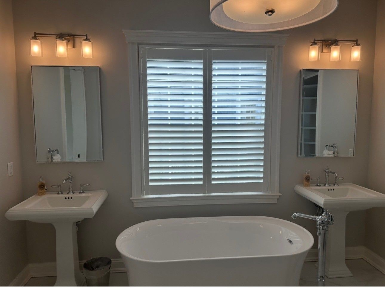Bathroom with a freestanding tub, two pedestal sinks, mirrors, and a window with shutters.