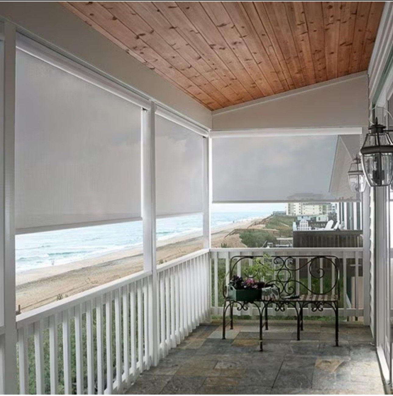 Balcony with white blinds overlooking a beach and ocean. Wooden ceiling, gray tile floor, and white railing.