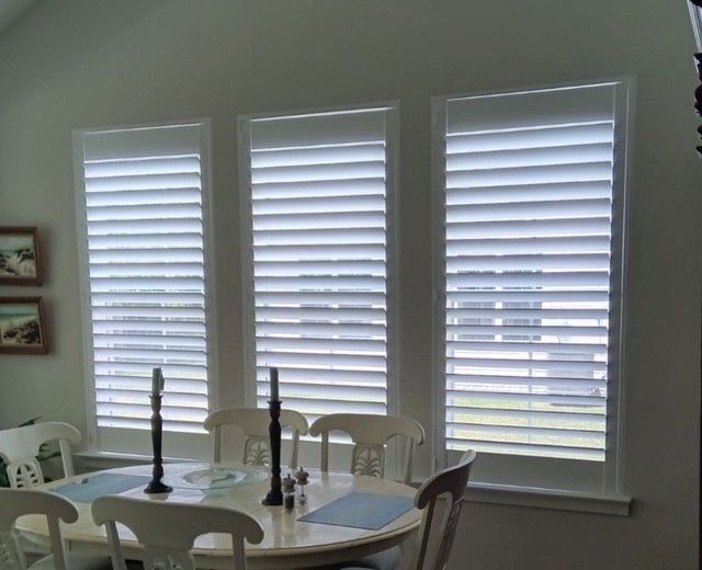 White slatted shutters on three windows above a dining table with white chairs.