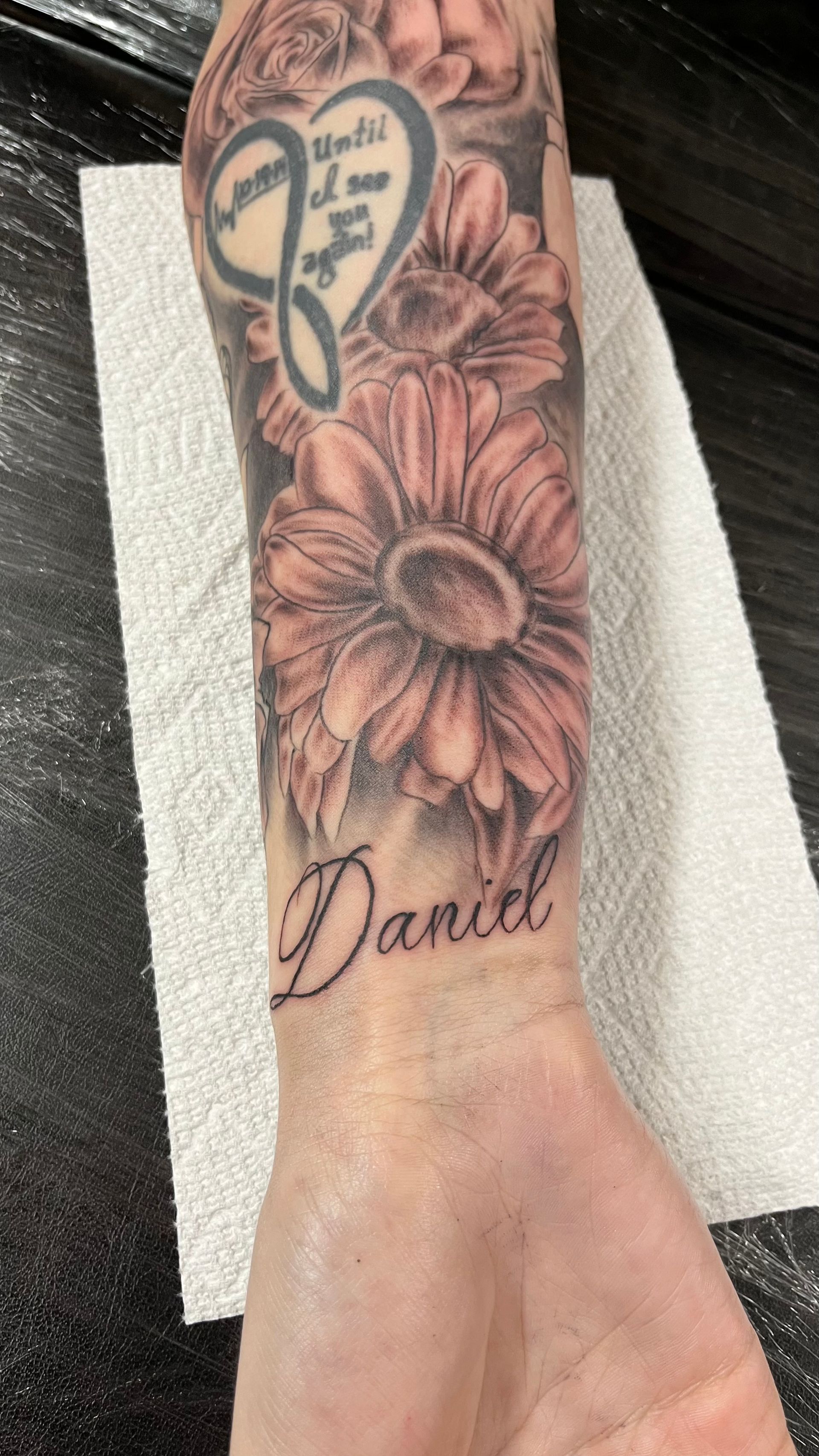 A person has a tattoo of flowers and the name daniel on their forearm.
