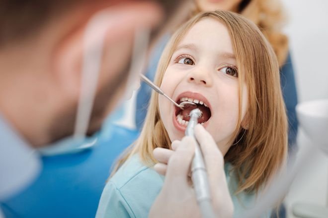 A dentist performs a dental procedure on a young patient in an office, using a handheld instrument inside their mouth.