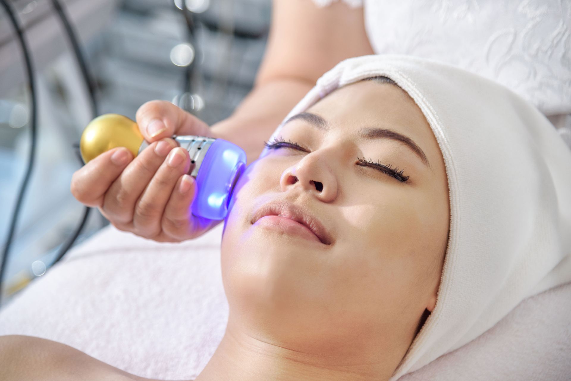 A person receives a blue light facial treatment with a handheld device while lying down in a spa setting.