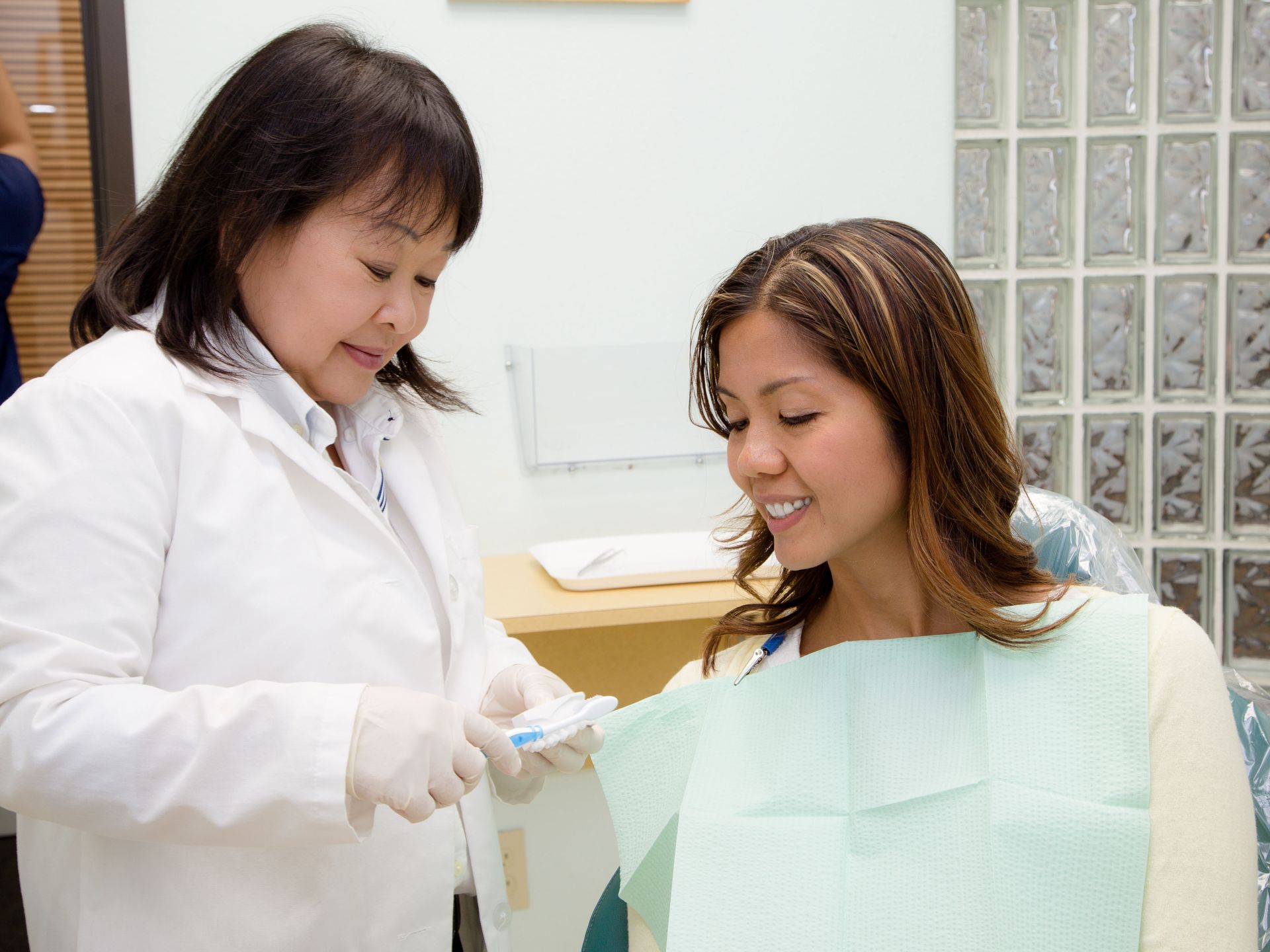 A dental professional in a white coat shows a toothbrush to a smiling patient wearing a dental bib.
