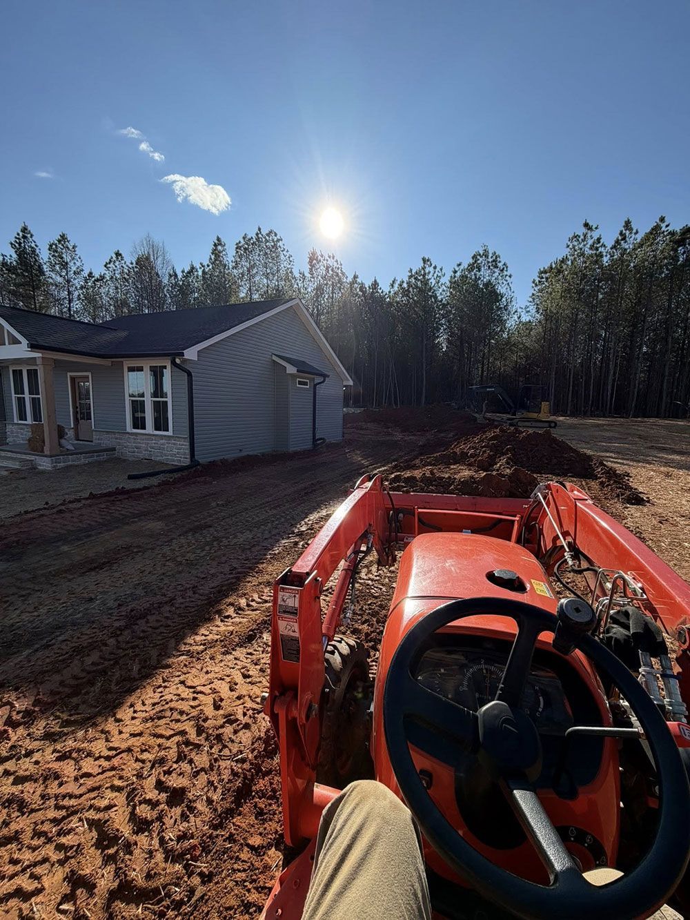 Orange tractor towing mud beside a house under bright sun, with trees in the background