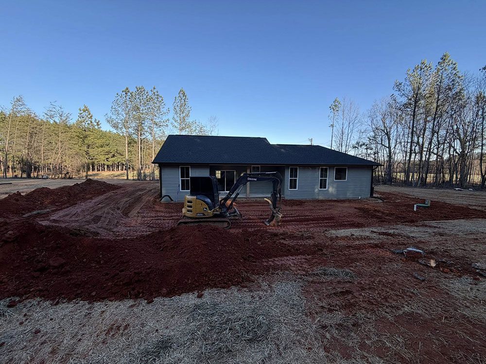 Single-story house under construction on a dirt lot with a front loader and pine trees in the background