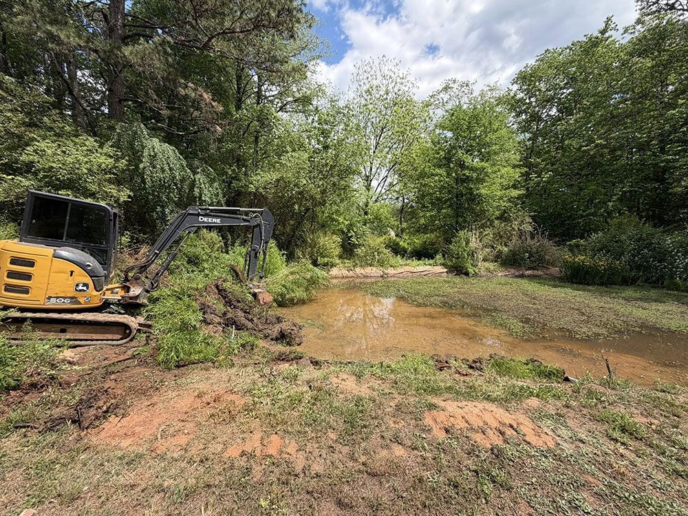 Yellow excavator clearing muddy ground beside a wooded pond