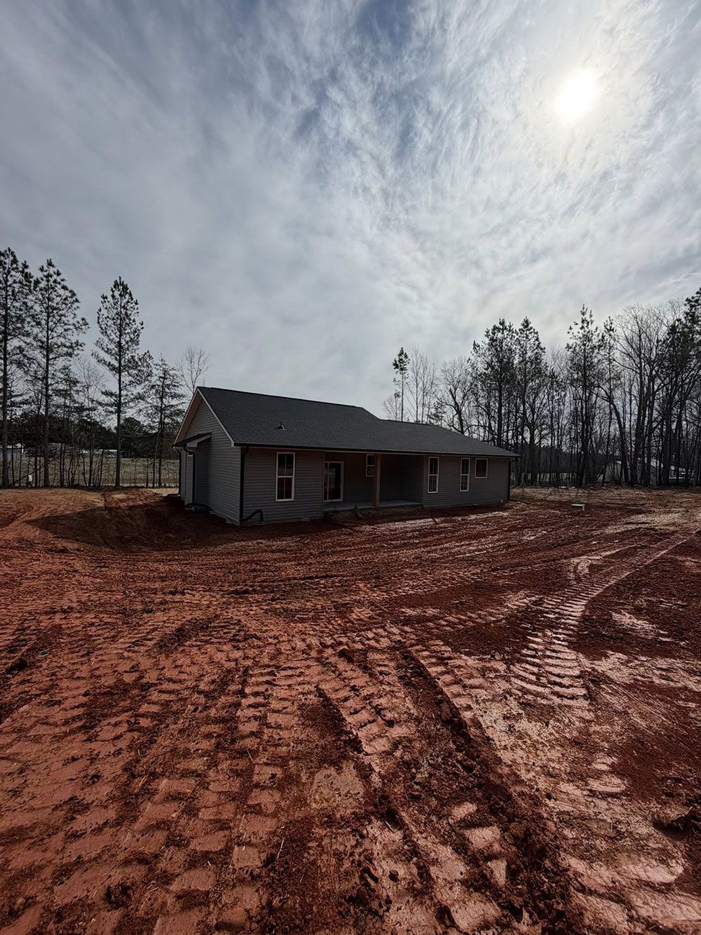 Single-story house under construction on a red dirt lot with tire tracks and cloudy sky