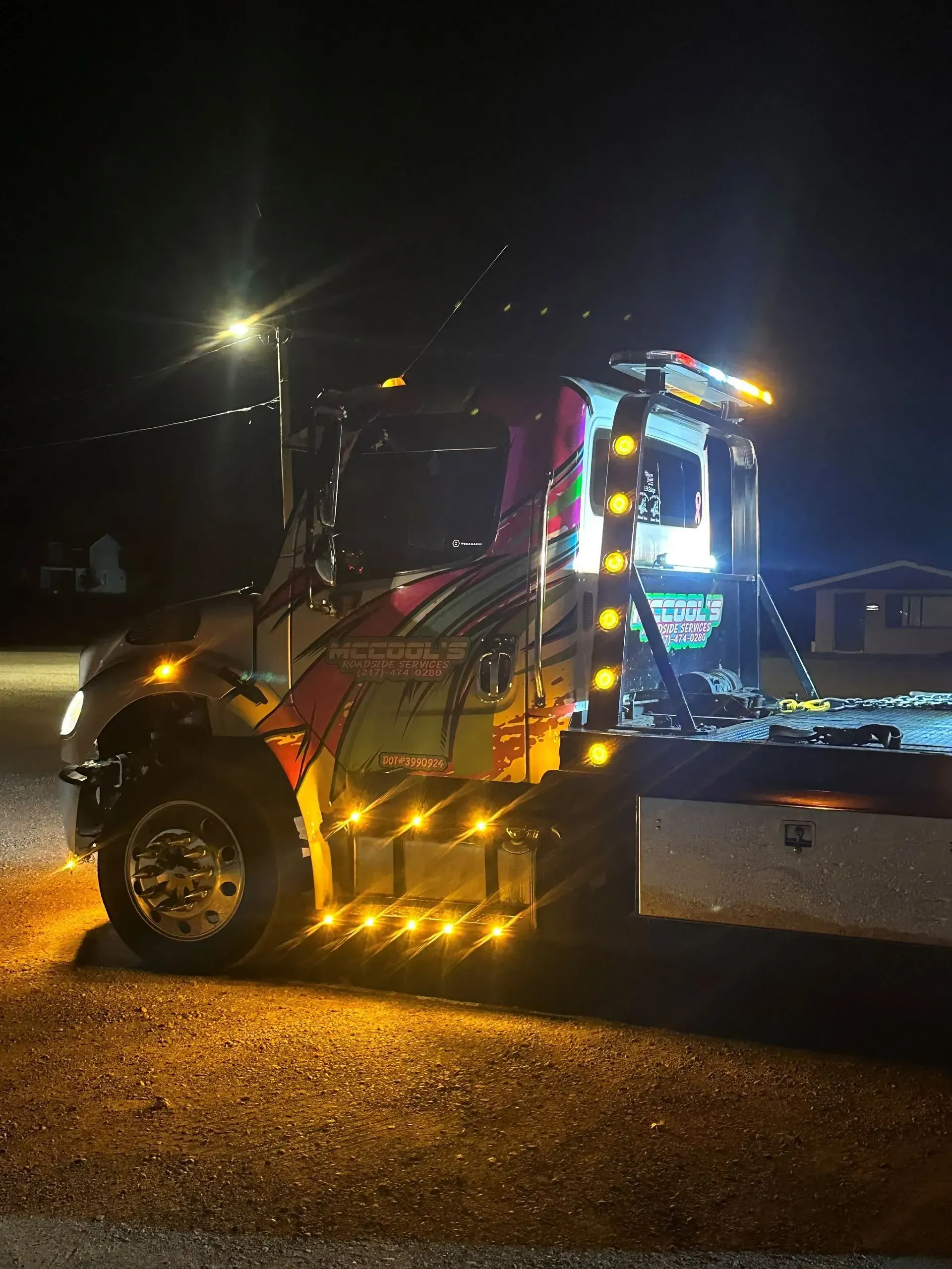 Brightly lit tow truck at night, amber lights flashing, parked on gravel.