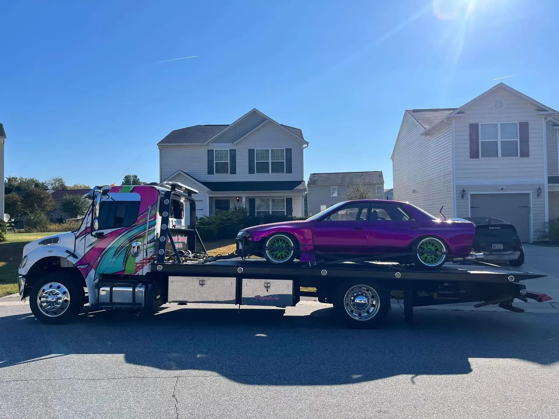 Purple car on tow truck in front of suburban houses on a sunny day.