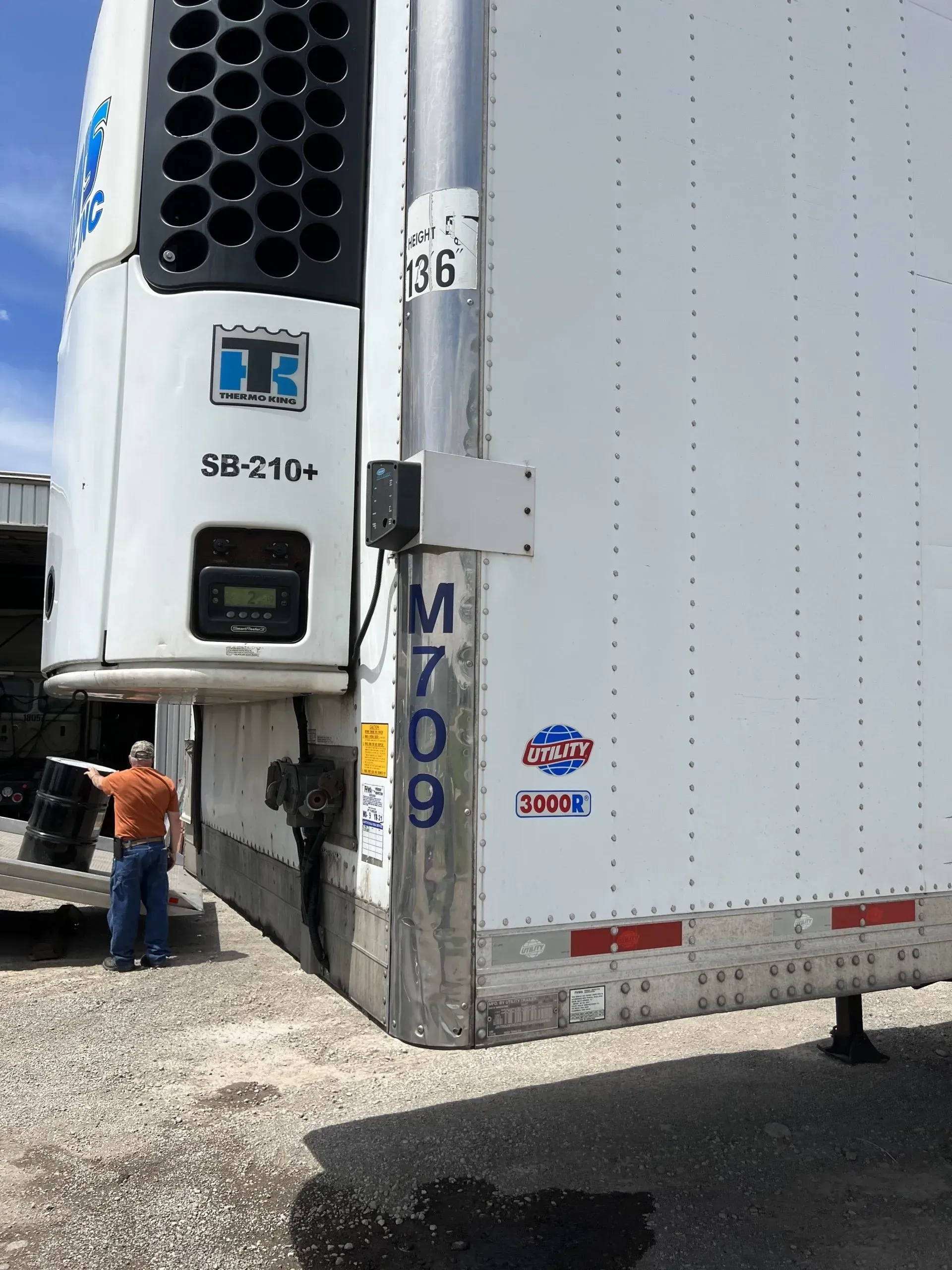 Refrigerated semi-trailer with a man near the front. White trailer, blue and black cooling unit. Outdoors.