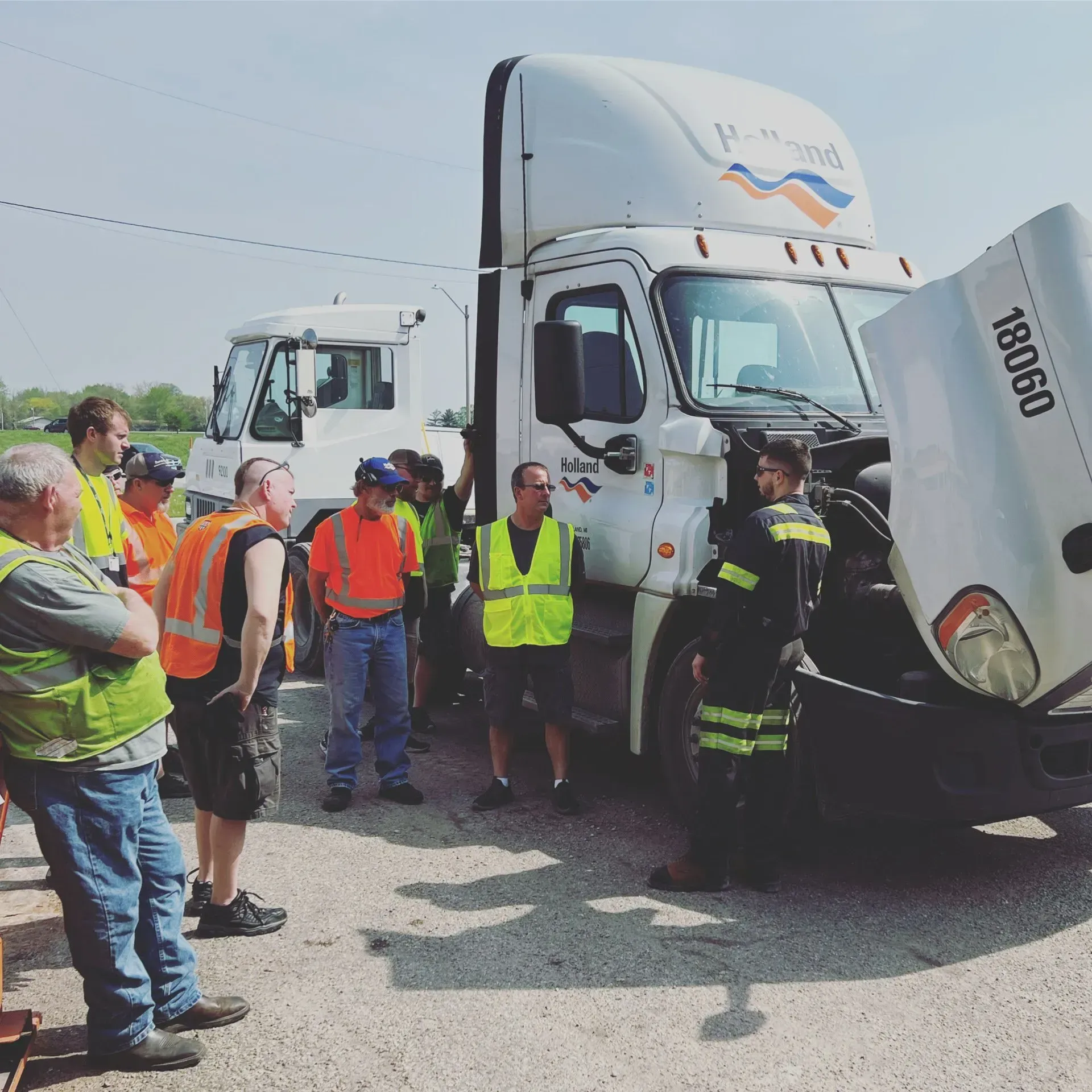 Group of people examining a semi-truck with the hood open, likely for maintenance or training outdoors.
