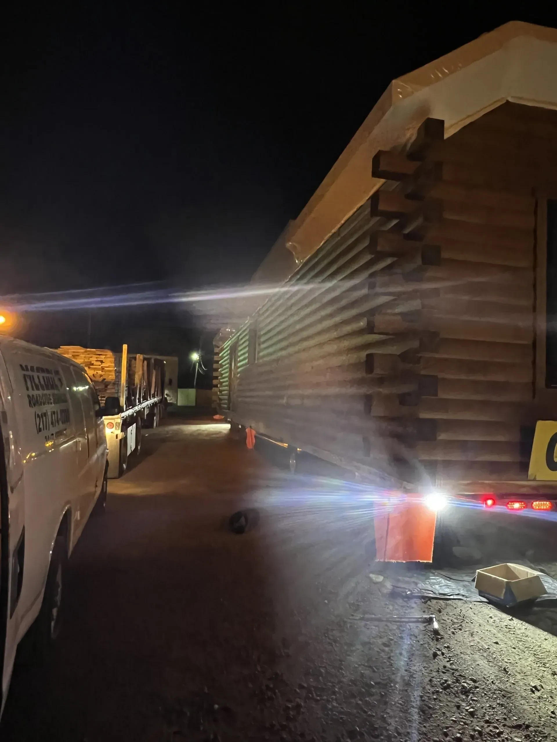 A log cabin on a trailer at night; side view. Headlights illuminate the logs, and there is a white van nearby.