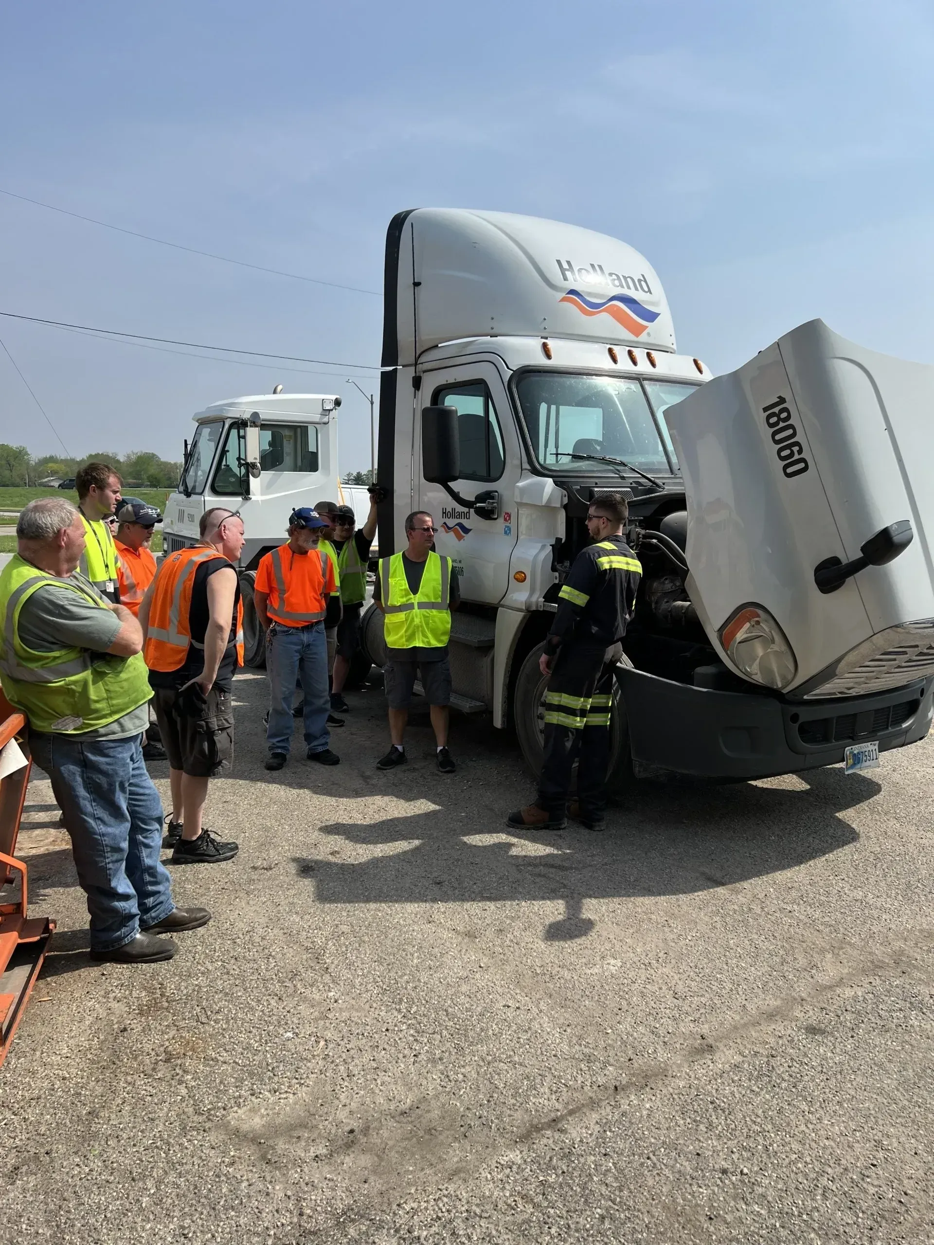 Group of people inspecting a semi-truck with open hood; several wear vests, under bright sky.