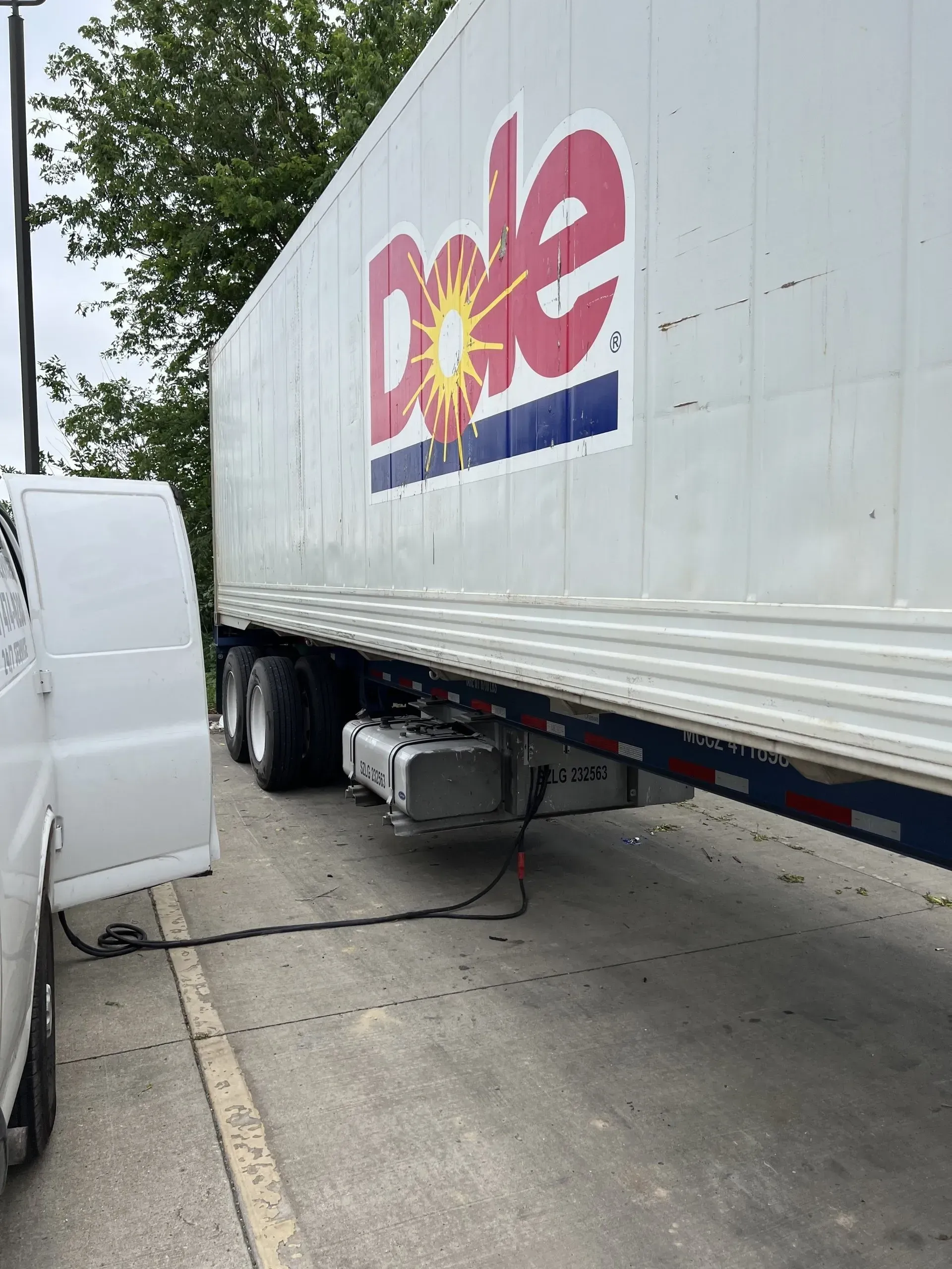 Dole semi-trailer next to a white van, connected by a black cable, parked on asphalt.