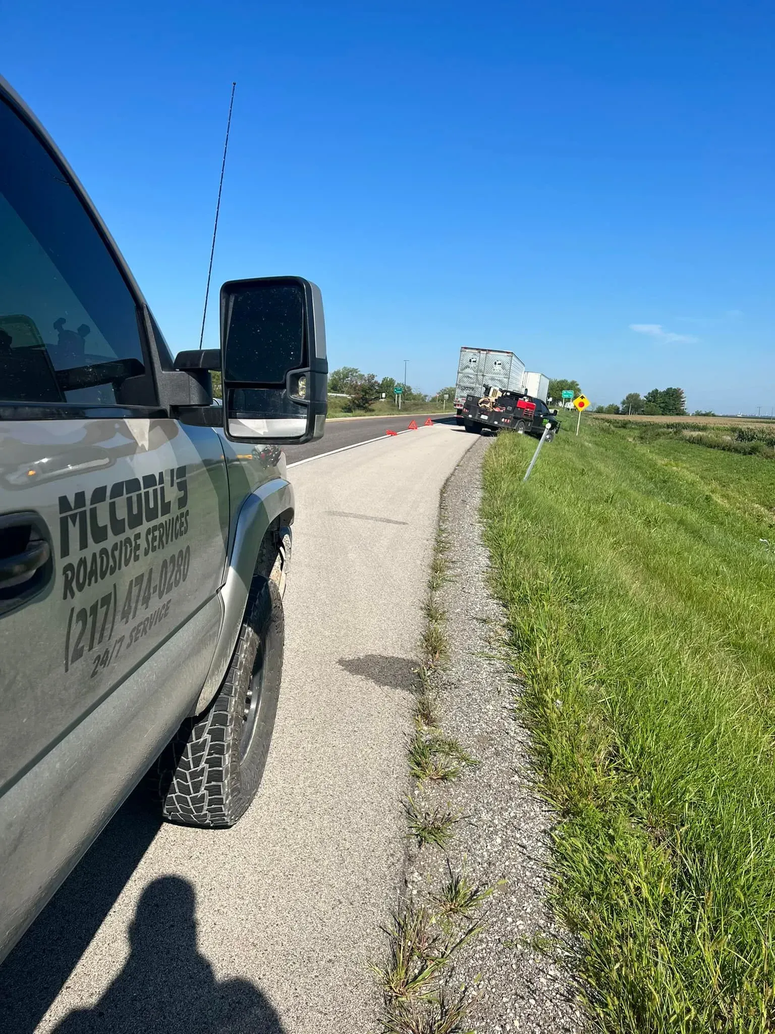 Truck on roadside. Another truck and vehicle in the distance. Green field on the right. Blue sky.