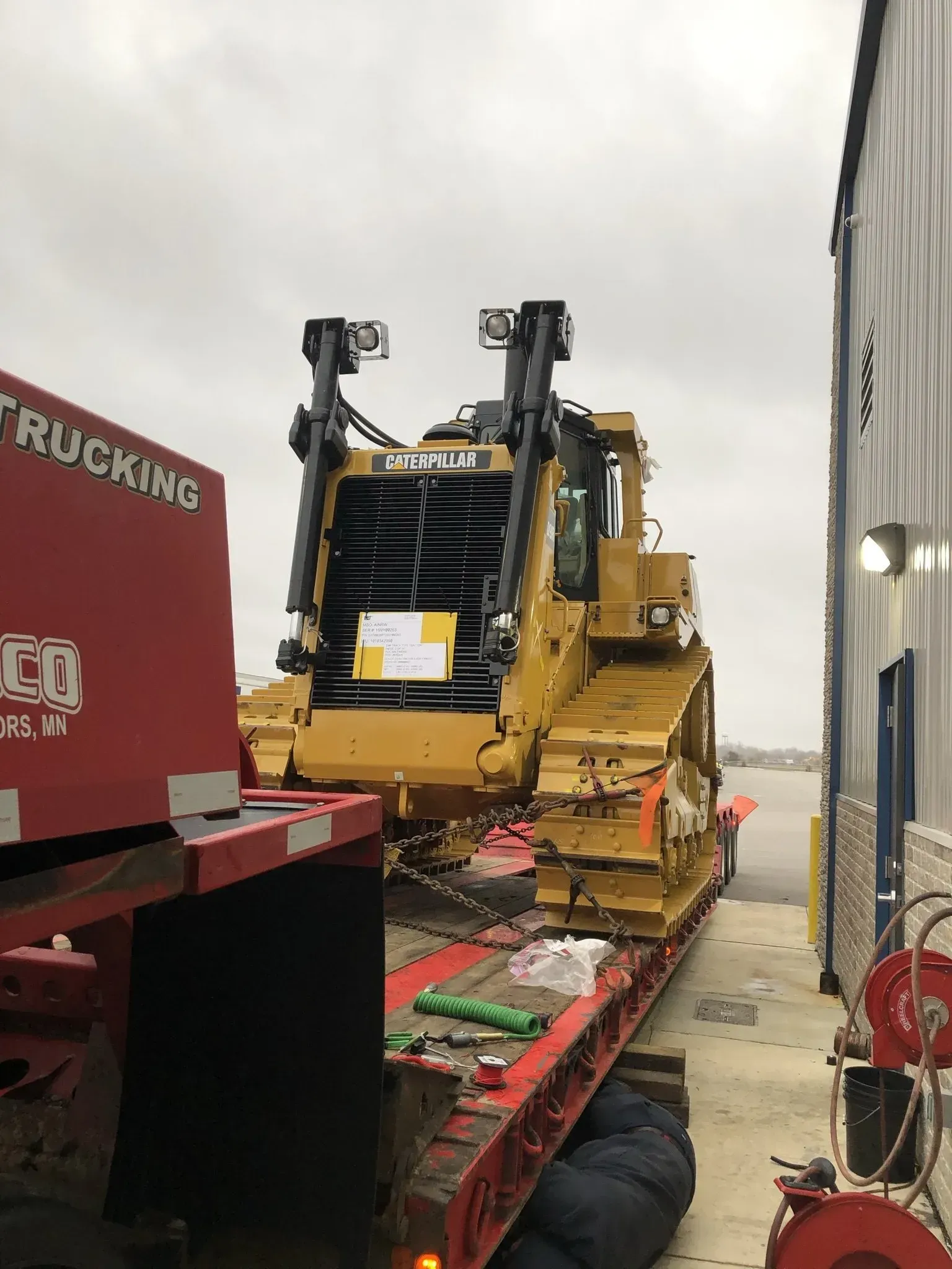 Yellow Caterpillar bulldozer loaded on a red flatbed truck, parked next to a building. Cloudy sky.