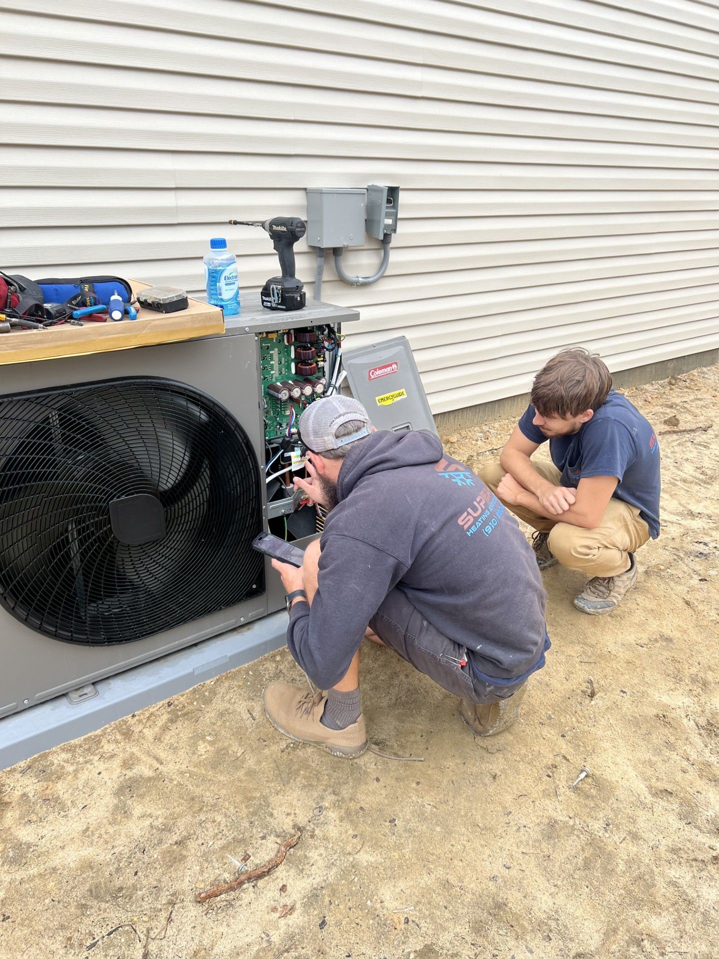 Two people work on a gray HVAC unit outside a building with light siding. One kneels and works on the wiring.