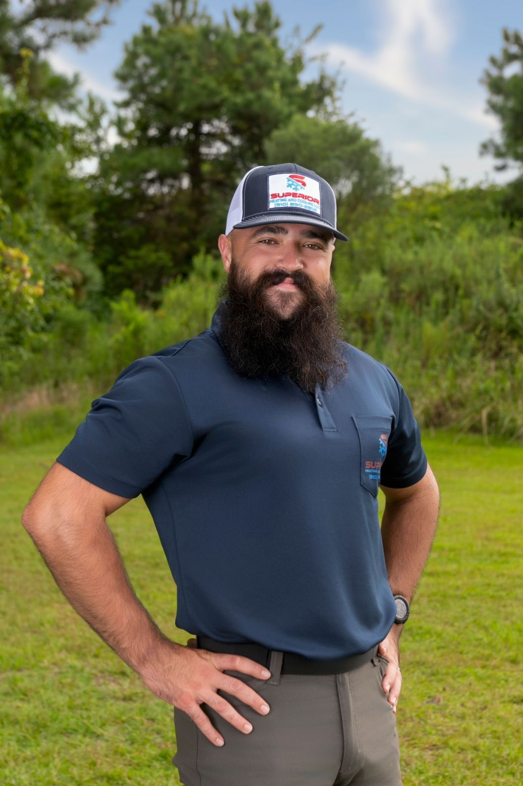 Man with long beard, wearing cap and polo shirt, stands with hands on hips in grassy outdoor setting.