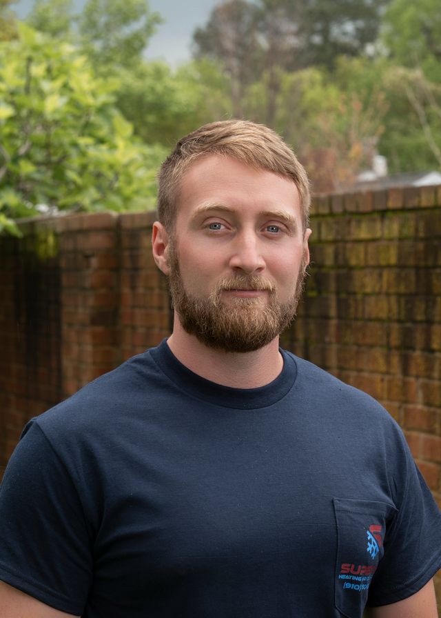 Man with a beard in a navy blue shirt in front of a brick wall and foliage.