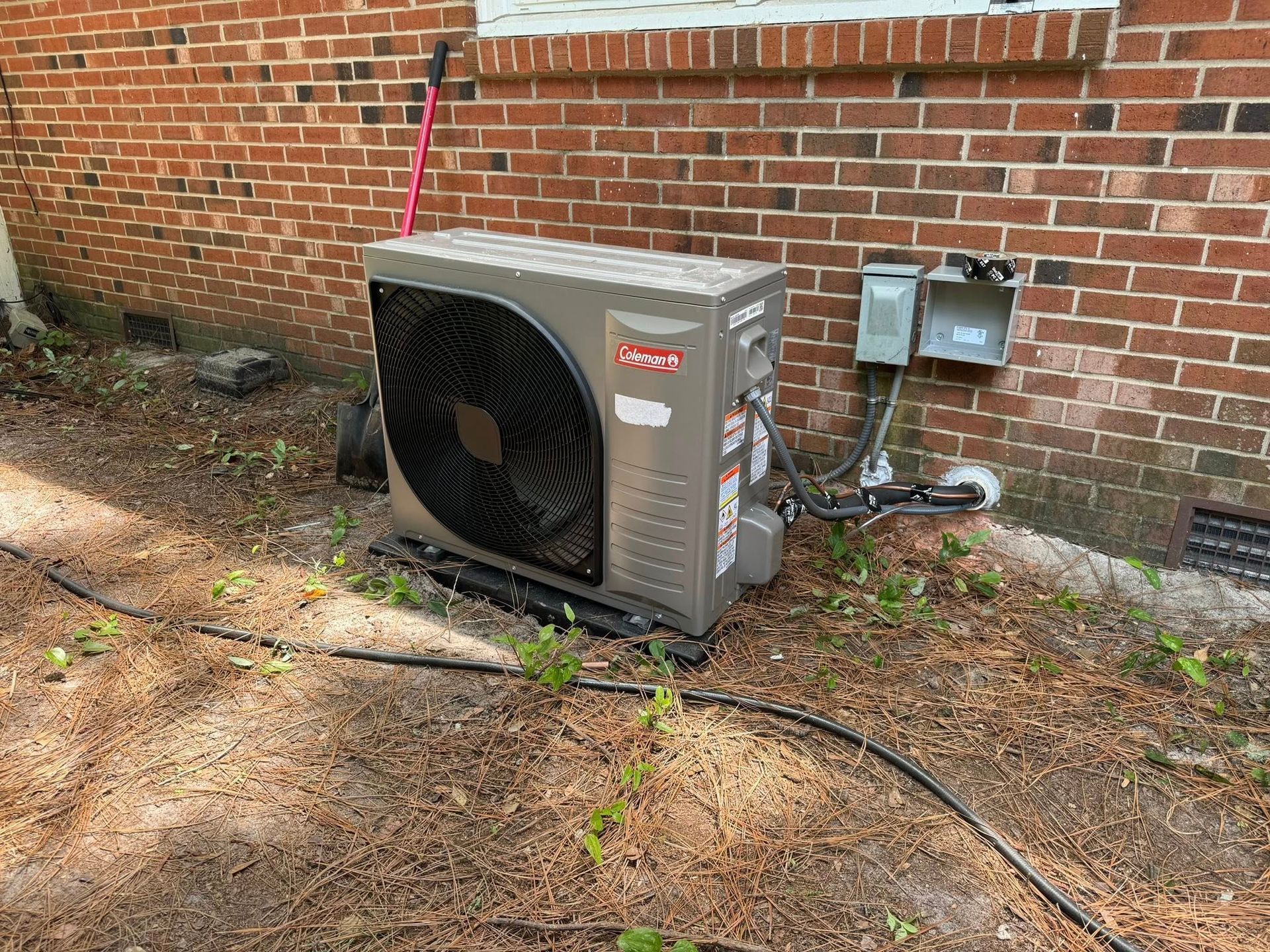 An outdoor air conditioning unit next to a brick wall. Black fan cover and electrical boxes.