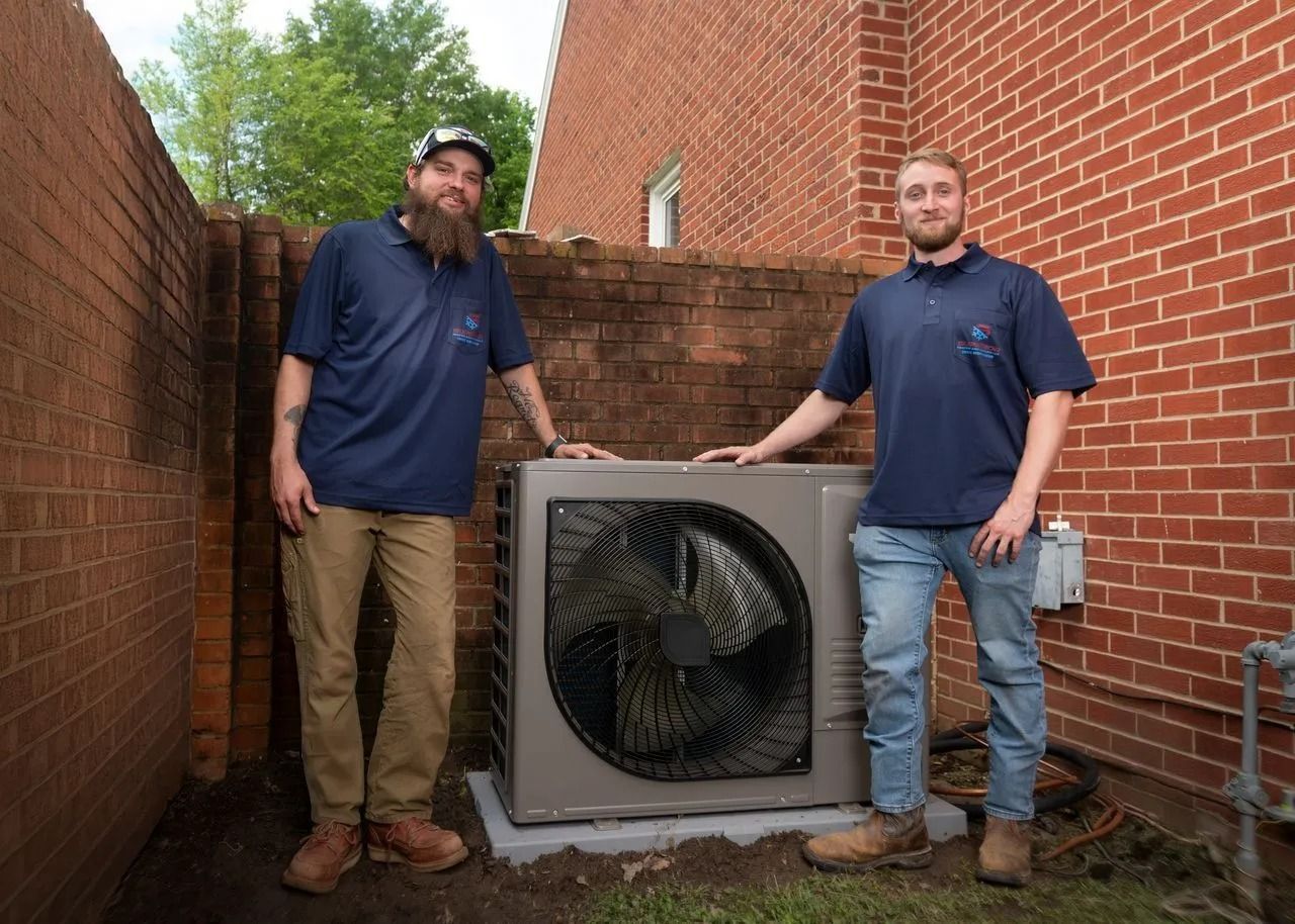 Two men stand beside an HVAC unit in a brick-walled enclosure. They wear matching blue shirts, and brown pants and jeans.