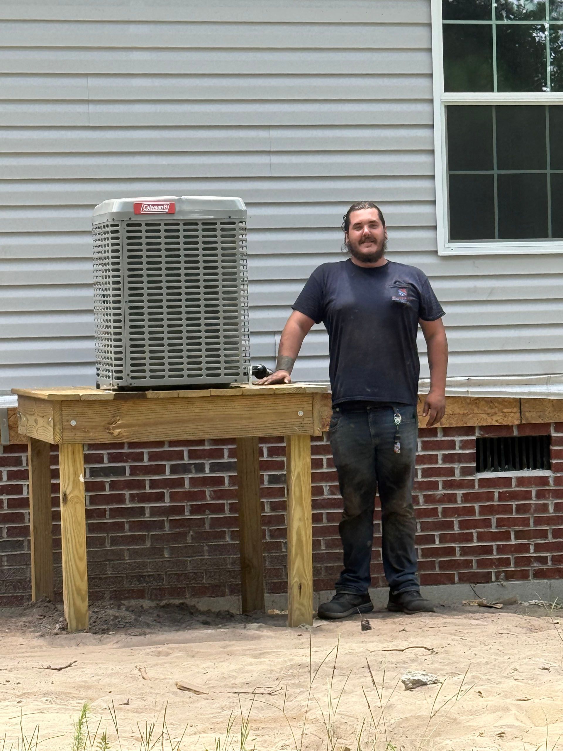 Man standing next to new air conditioning unit on wooden platform against a building with brick foundation.