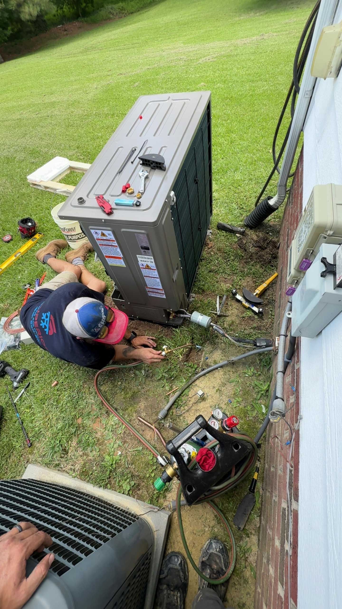 HVAC technician works on an air conditioning unit outside a brick building, with tools scattered around.