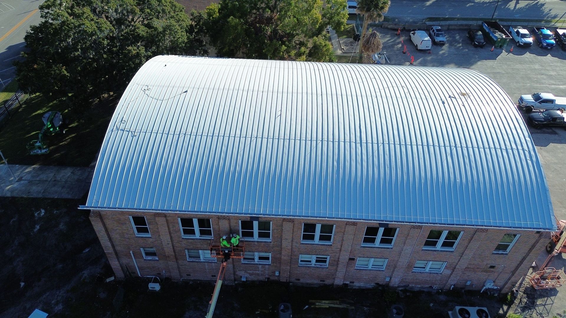 Aerial view of a brick building with a curved silver roof, surrounded by trees and parked cars.