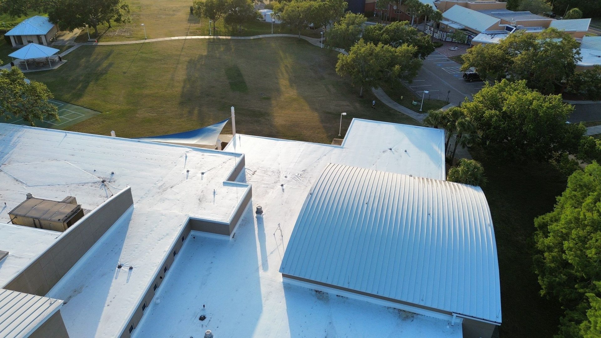 Aerial view of a white-roofed building beside a grassy yard and trees in bright daylight
