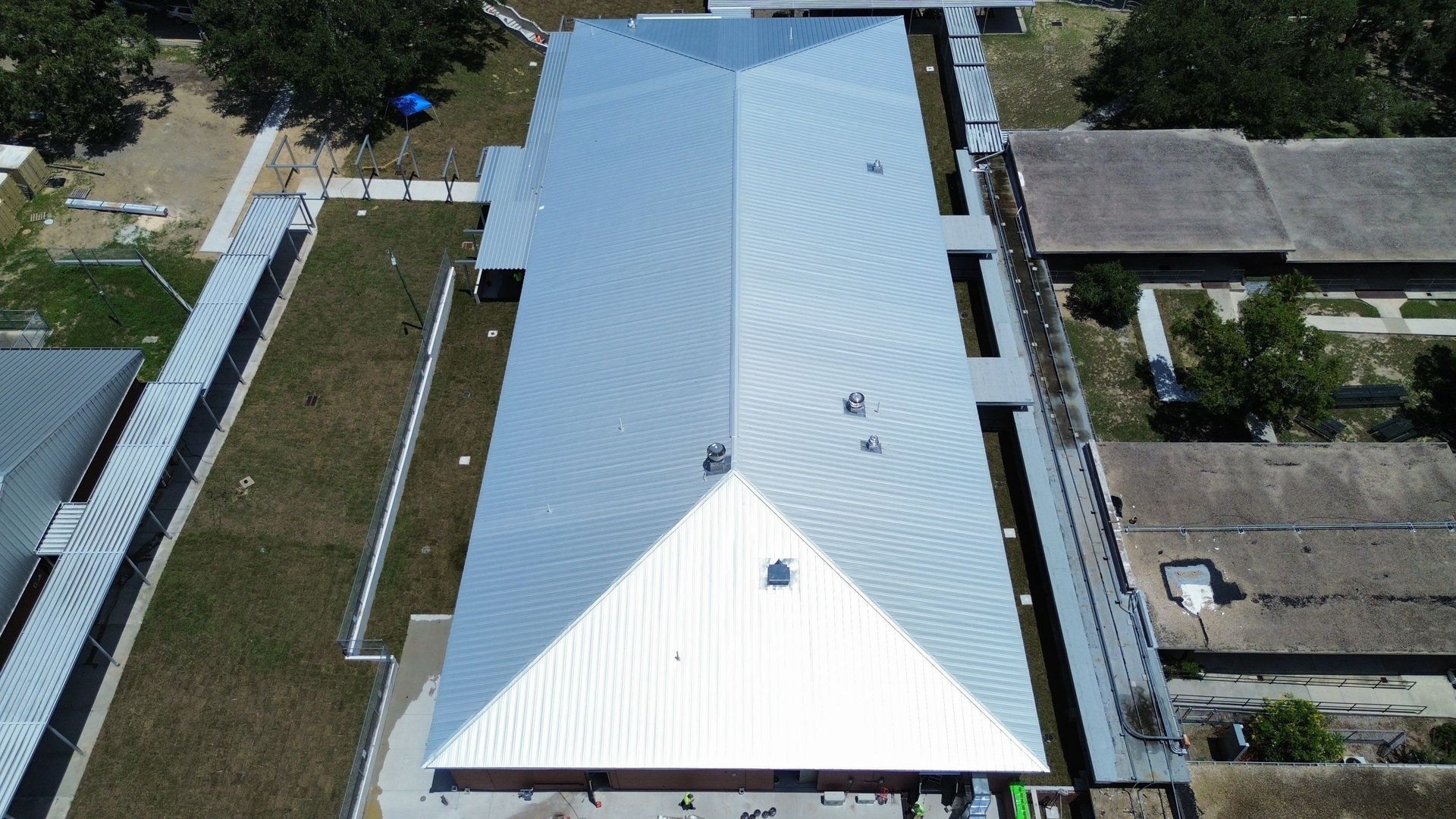 Aerial view of a long white-roofed building with side yards and neighboring homes.
