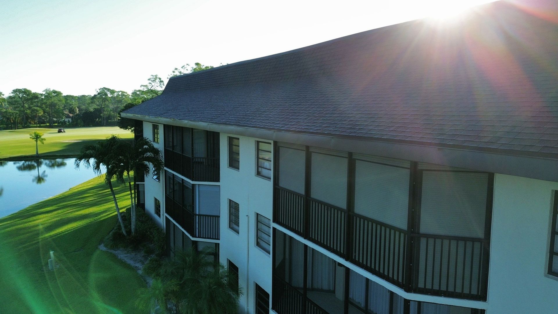 Sunlit lakeside house with a dark roof and screened porch beside green lawn and water