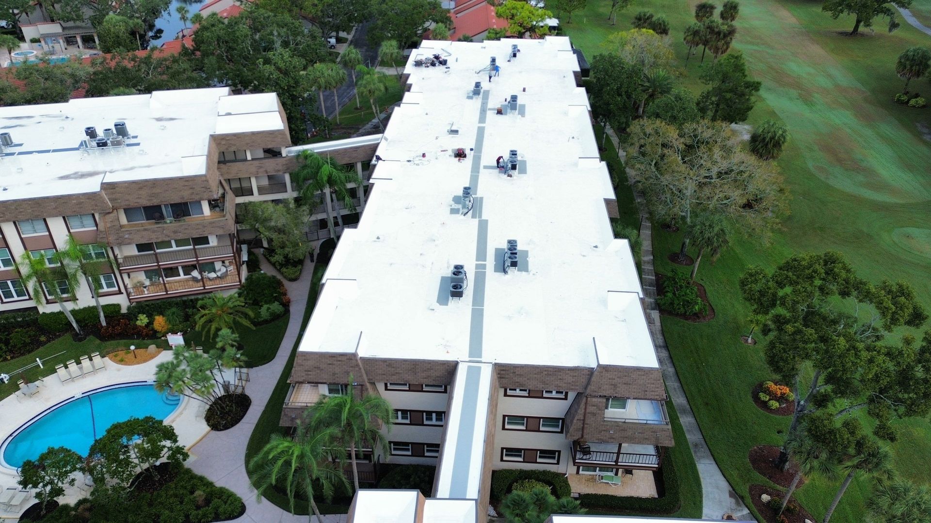 Aerial view of a white-roofed apartment complex beside a swimming pool and green lawn.