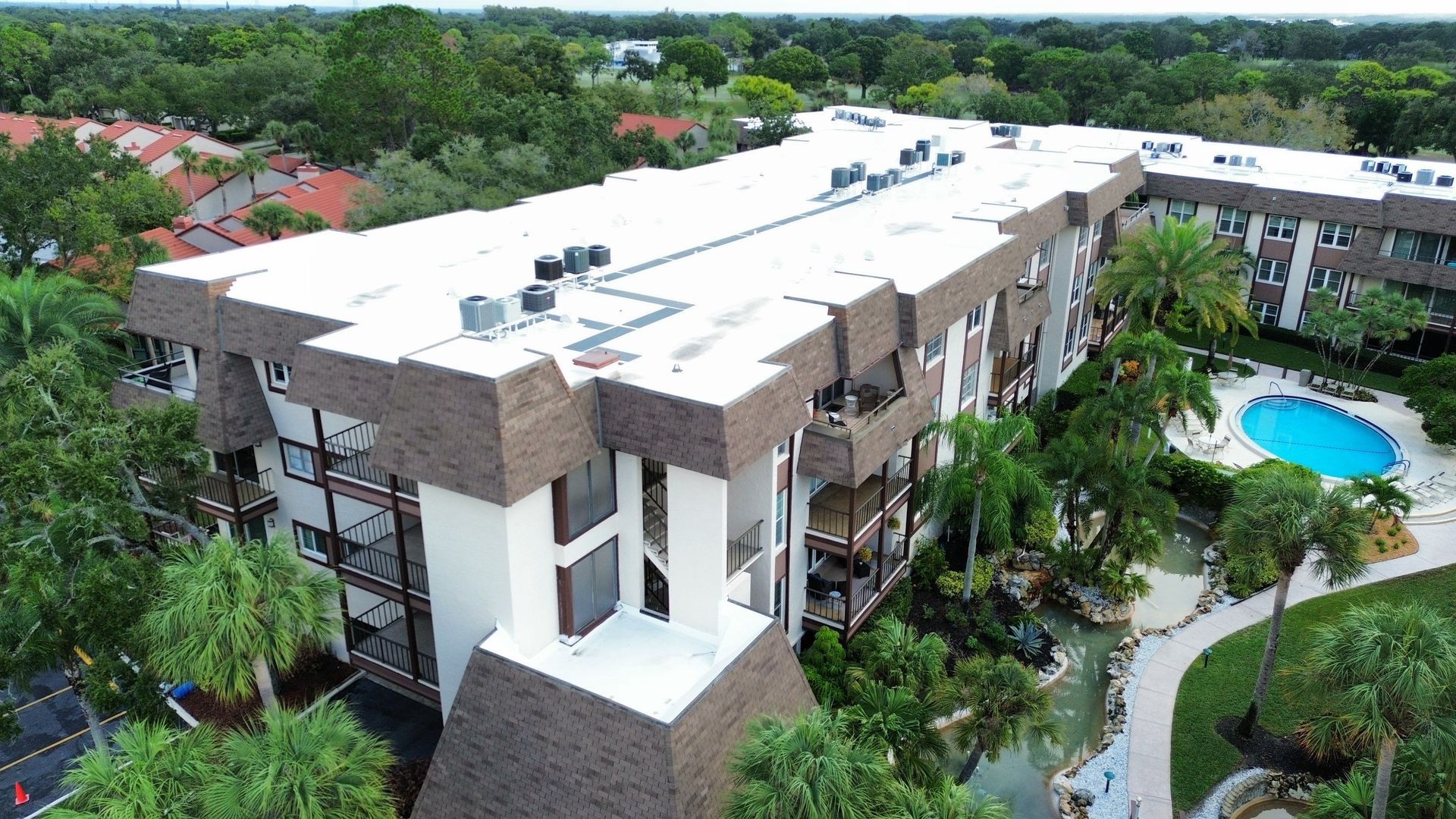 Aerial view of a white-roofed apartment complex with palm trees and a blue swimming pool.
