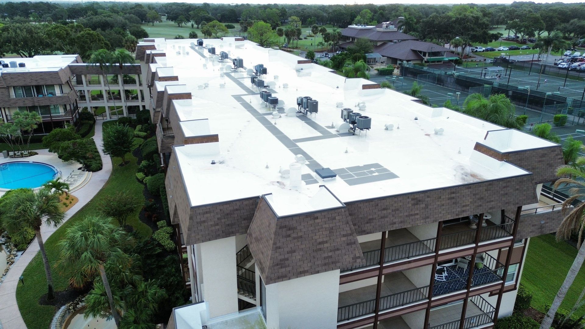 Aerial view of a white-roofed resort building beside a pool and surrounded by lush greenery.