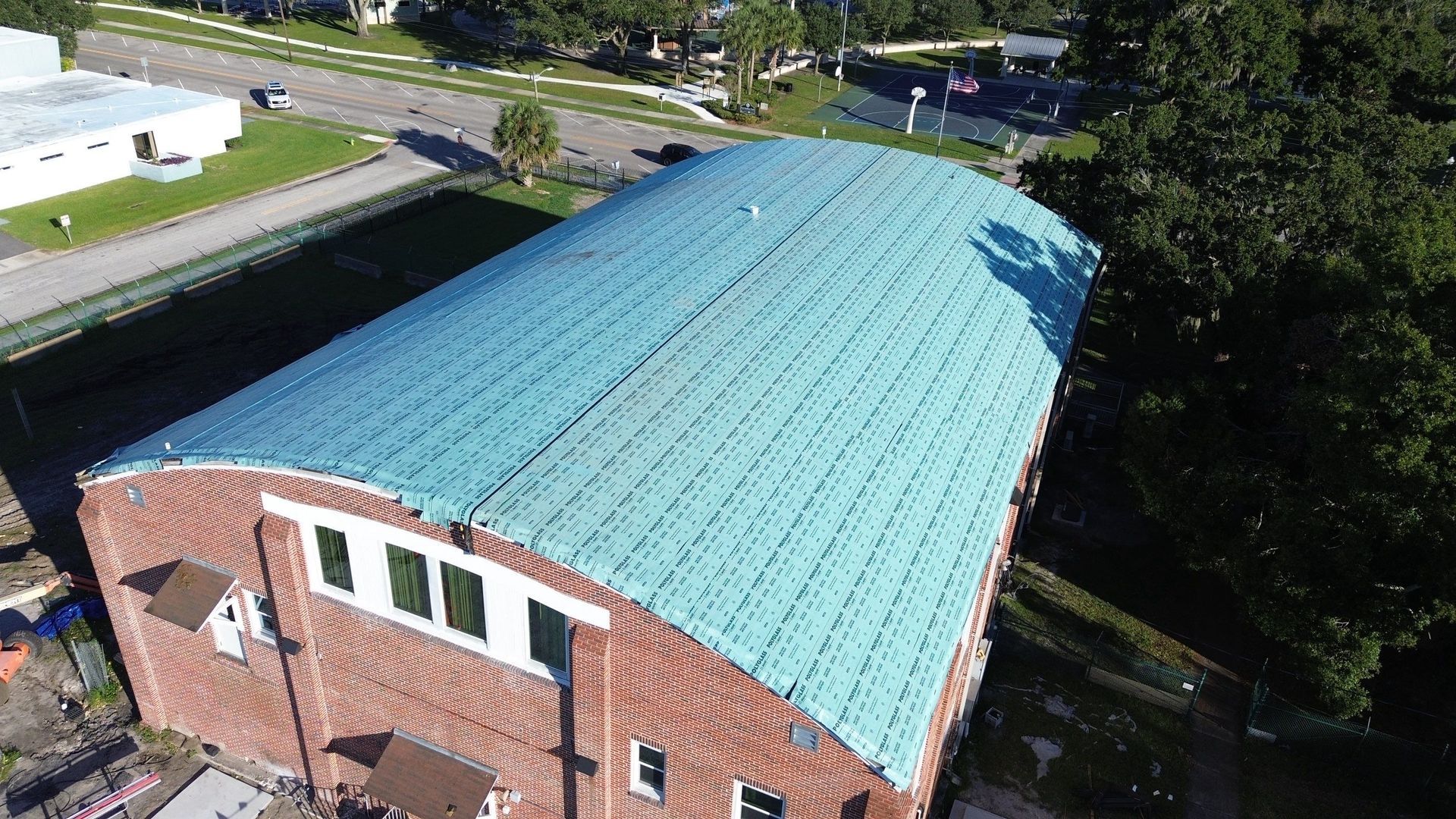 Aerial view of a brick building with a turquoise roof beside a road and trees.